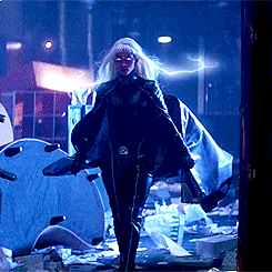 A Woman In A Cape Is Standing In A Dark Room With A Lightning Bolt Behind Her GIF