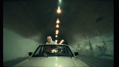A Woman In A Car Is Driving Through A Tunnel With Lights Coming Out Of The Ceiling GIF