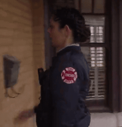 A Woman In A Chicago Fire Department Jacket Is Standing In Front Of A Window GIF