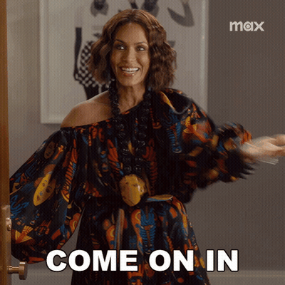 A Woman In A Colorful Dress Is Standing In Front Of A Door And Says Come On In GIF
