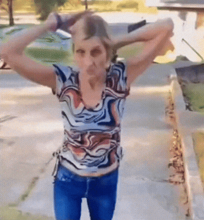 A Woman In A Colorful Shirt Is Standing On A Sidewalk With Her Hands In Her Hair . GIF