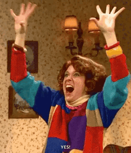 A Woman In A Colorful Sweater Is Raising Her Hands In The Air GIF