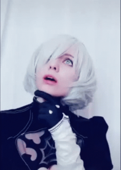 A Woman In A Cosplay Costume With White Hair And Black Gloves Is Standing In Front Of A White Wall . GIF