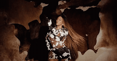 A Woman In A Cow Print Dress Is Standing In Front Of A Cow Skin Rug . GIF