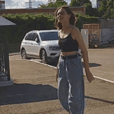 A Woman In A Crop Top And Jeans Is Standing In A Parking Lot . GIF
