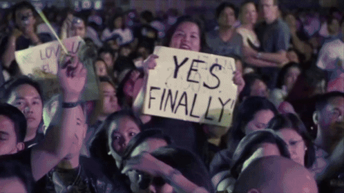 A Woman In A Crowd Holds Up A Sign That Says " Yes Finally " GIF