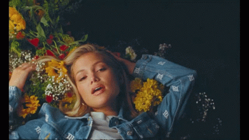 A Woman In A Denim Jacket Is Laying In A Bed With Flowers GIF