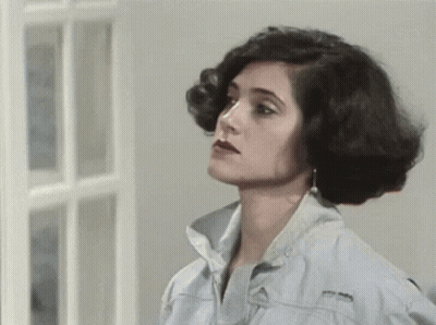 A Woman In A Denim Jacket Is Standing In Front Of A Window And Looking At The Camera . GIF