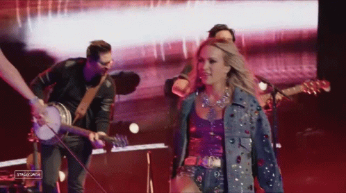 A Woman In A Denim Jacket Is Walking On Stage With A Band GIF