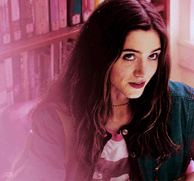 A Woman In A Denim Jacket Stands In Front Of A Bookshelf And Looks At The Camera GIF
