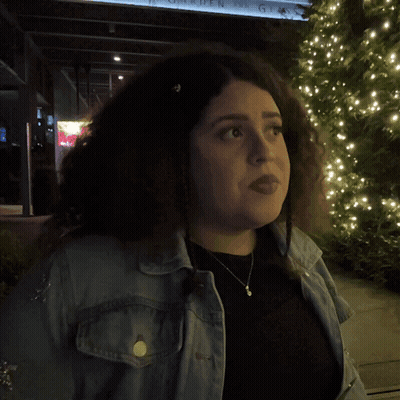 A Woman In A Denim Jacket Stands In Front Of A Sign That Says What GIF