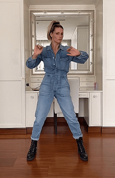 A Woman In A Denim Jumpsuit And Black Boots Is Dancing In Front Of A Mirror GIF