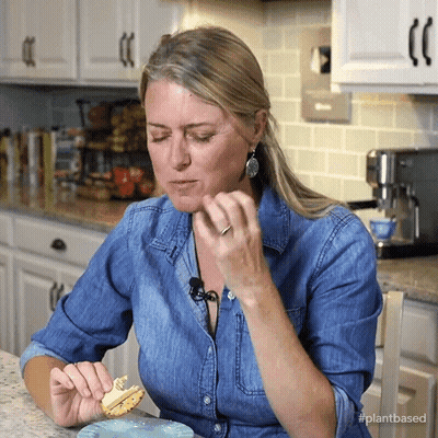 A Woman In A Denim Shirt Is Eating A Sandwich In A Kitchen With The Hashtag Plantbased GIF