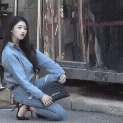 A Woman In A Denim Shirt Is Kneeling Down On The Sidewalk Holding A Black Purse That Says Jil Sander On It GIF