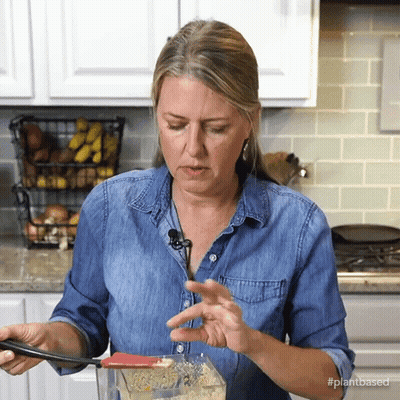 A Woman In A Denim Shirt Is Stirring Something In A Blender With The Hashtag #Plantbased On The Bottom GIF