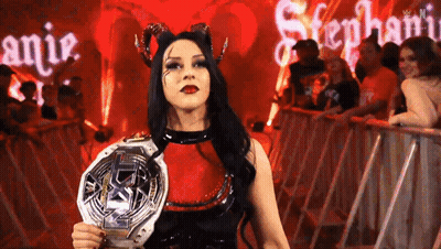 A Woman In A Devil Costume Is Holding A Championship Belt In Front Of A Crowd . GIF