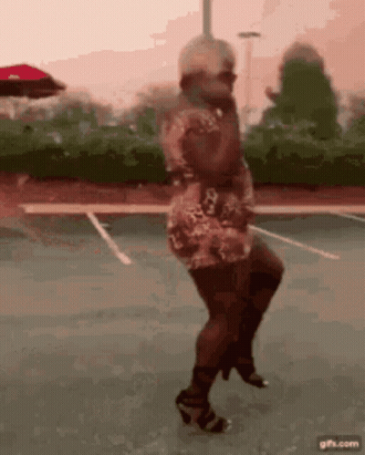 A Woman In A Dress And Heels Is Dancing On A Parking Lot . GIF