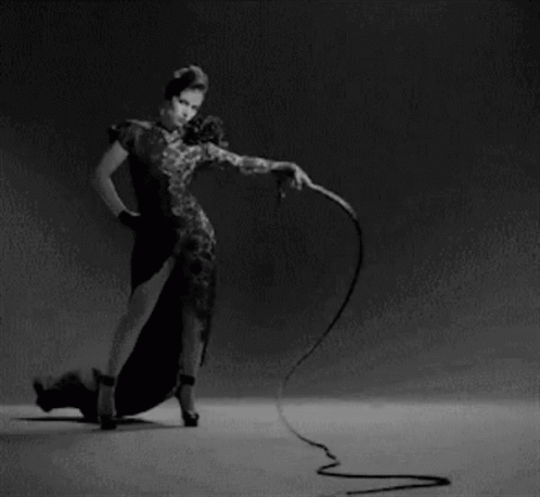 A Woman In A Dress Is Holding A Whip In Her Hand . GIF
