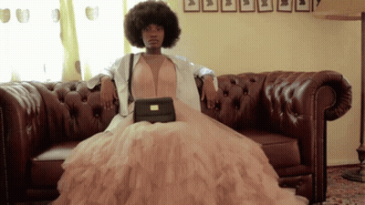 A Woman In A Dress Is Sitting On A Couch Holding A Black Purse GIF