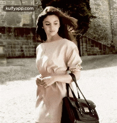 A Woman In A Dress Is Walking Down A Street Holding A Black Bag . GIF