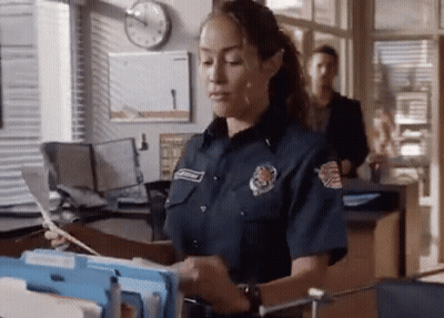 A Woman In A Firefighter 'S Uniform Is Standing In An Office Looking At A Stack Of Papers . GIF