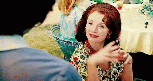 A Woman In A Floral Dress Is Sitting At A Table With A Man And Waving At Him . GIF