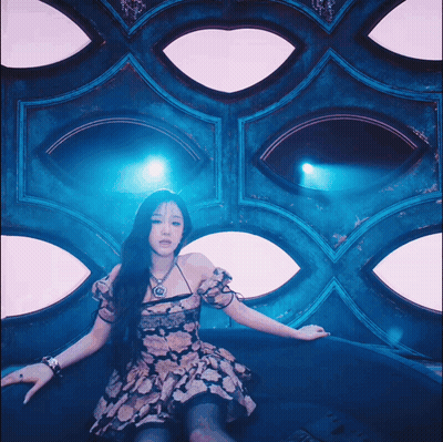 A Woman In A Floral Dress Is Sitting In Front Of A Wall With Eyes GIF