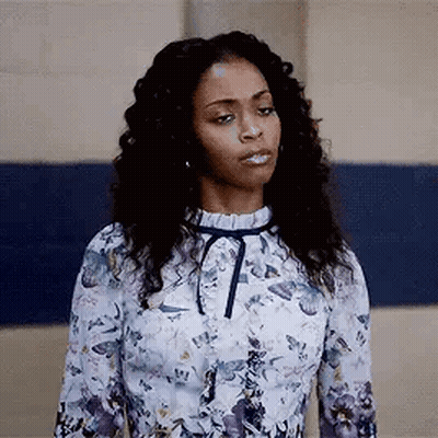 A Woman In A Floral Dress Is Standing In Front Of A Blue Wall And Making A Funny Face . GIF