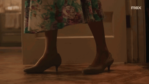 A Woman In A Floral Dress Is Standing In Front Of A Door With The Word Max On The Bottom Right GIF