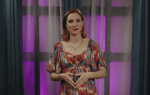 A Woman In A Floral Dress Is Standing In Front Of A Window With Purple Curtains GIF