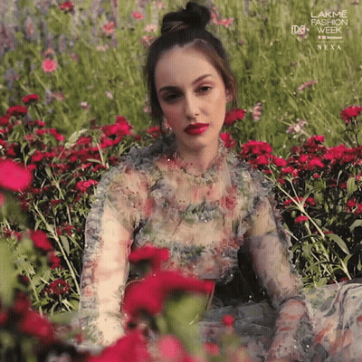 A Woman In A Floral Dress Sits In A Field Of Flowers With A Lakme Fashion Week Next Advertisement Behind Her GIF