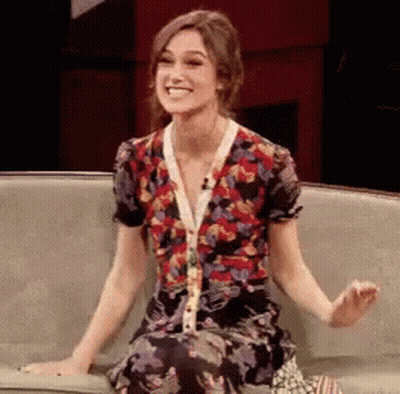 A Woman In A Floral Dress Sits On A Couch Smiling GIF