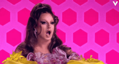 A Woman In A Floral Dress Stands In Front Of A Pink Background With A Letter V On It GIF