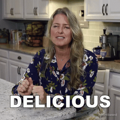 A Woman In A Floral Shirt Is Sitting At A Kitchen Counter And The Word Delicious Is Above Her GIF