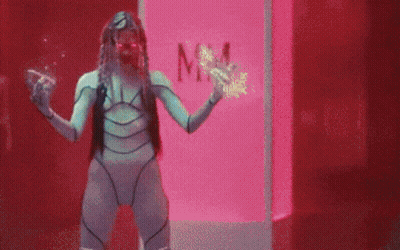 A Woman In A Futuristic Costume Is Standing In Front Of A Pink Box With The Word Mm On It . GIF