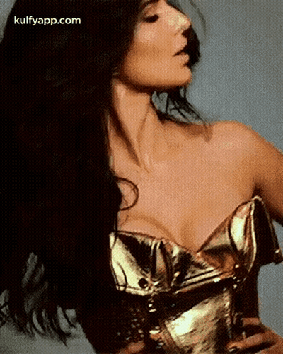 A Woman In A Gold Corset With Her Hair Blowing In The Wind Is Looking At The Camera . GIF