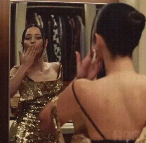A Woman In A Gold Dress Is Looking At Herself In The Mirror GIF