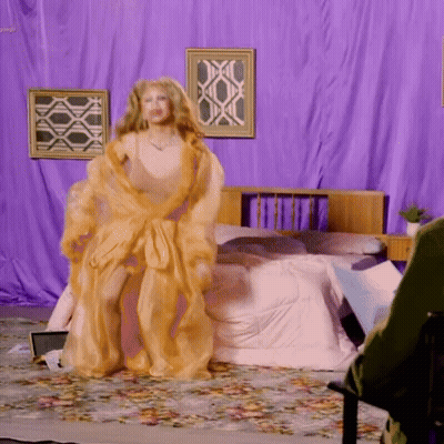 A Woman In A Gold Robe Is Standing In A Bedroom With Purple Curtains GIF