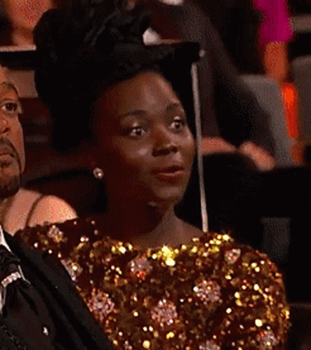 A Woman In A Gold Sequined Top Is Sitting In A Dark Room GIF