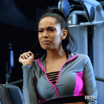 A Woman In A Gray And Pink Sports Bra Is Sitting In A Gym Making A Funny Face GIF