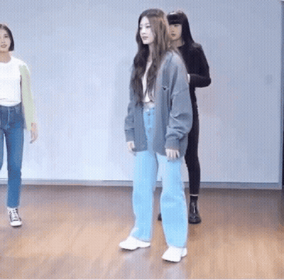 A Woman In A Gray Cardigan And Blue Jeans Is Standing On A Wooden Floor In A Dance Studio . GIF