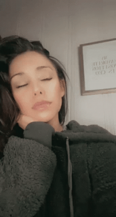 A Woman In A Gray Hoodie Is Sleeping With Her Eyes Closed And Her Hand On Her Face . GIF