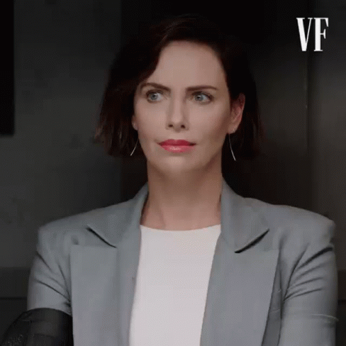 A Woman In A Gray Jacket And White Shirt Is Sitting In A Dark Room . GIF