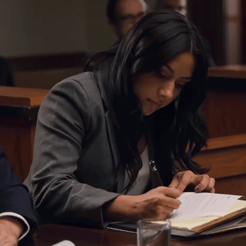 A Woman In A Gray Jacket Is Sitting At A Table Writing On A Piece Of Paper GIF