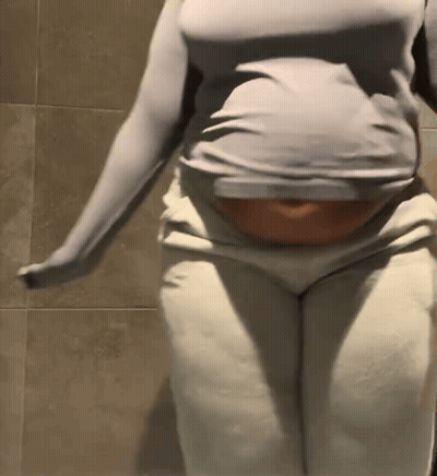 A Woman In A Gray Shirt And White Pants Is Standing In Front Of A Tiled Wall . GIF