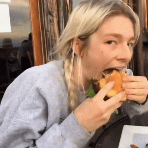A Woman In A Gray Sweatshirt Is Eating A Sandwich GIF
