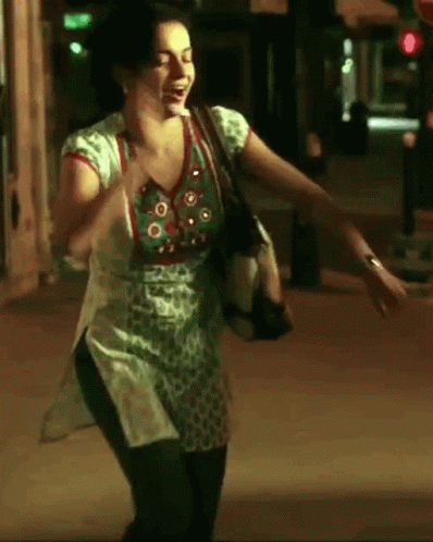 A Woman In A Green And White Top Is Dancing On A Street At Night GIF
