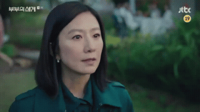 A Woman In A Green Coat Is Sitting In A Park Looking At The Camera . GIF