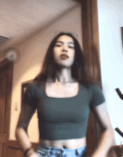 A Woman In A Green Crop Top And Shorts Is Standing In A Room . GIF