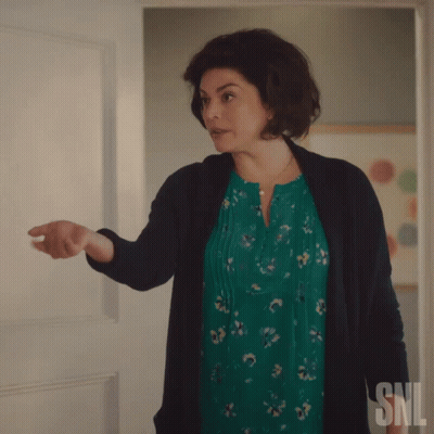 A Woman In A Green Dress And Black Cardigan Stands In Front Of A Snl Sign GIF